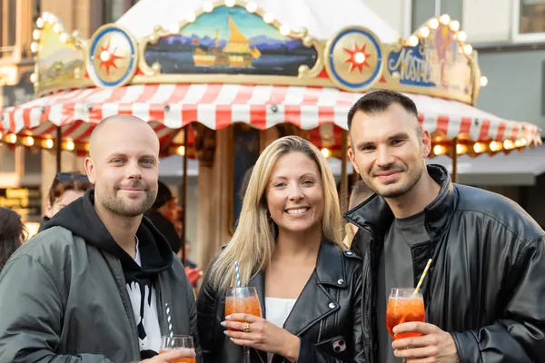 Drei Gäste mit Spritz vor einem Karussell mit rot-weißem Dach