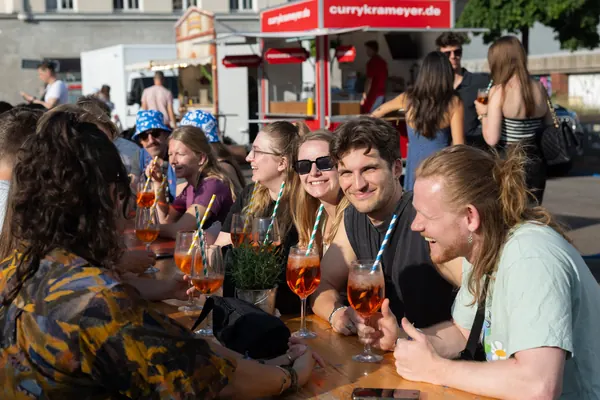Eine Gruppe mit Spritz an einem Tisch, dahinter ein Currywurst-Stand