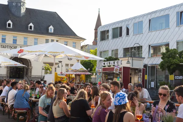 Gäste an Biertischen unter Schirmen vor dem Neptunbad